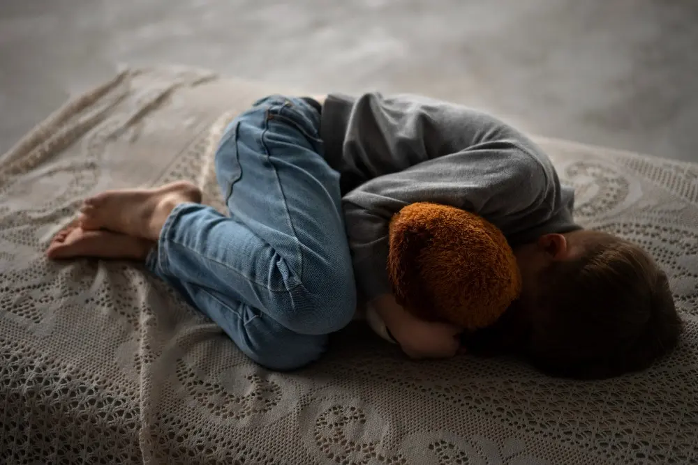 Toddler lying curled up on a bed holding a teddy bear, showing shyness or separation anxiety