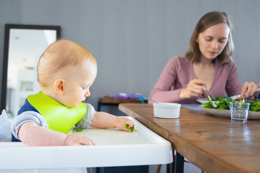 Baby in a high chair exploring food while mother eats at the table.