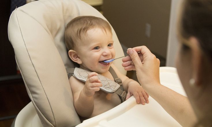 Smiling baby sitting in high chair being fed with a spoon.