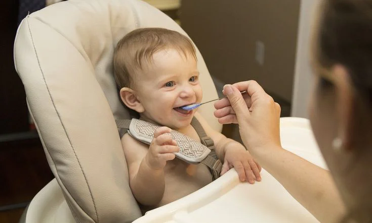 Signs Your Baby Is Ready for Solids (Not Just Age) 4 Smiling baby sitting in high chair being fed with a spoon.