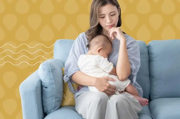 Mother sitting on a blue sofa holding a sleeping baby while looking worried.