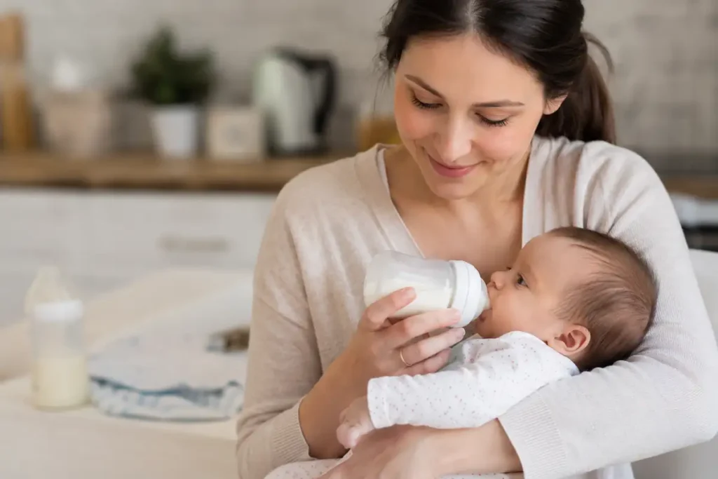 Mother feeding newborn baby with bottle in a warm cozy home kitchen, symbolizing safe formula feeding and bonding between parent and infant.